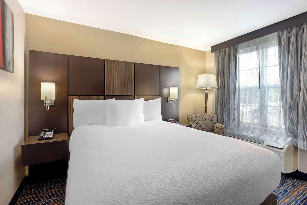 A hotel room with a large bed, a bedside table with a phone, a chair, and a window with curtains.