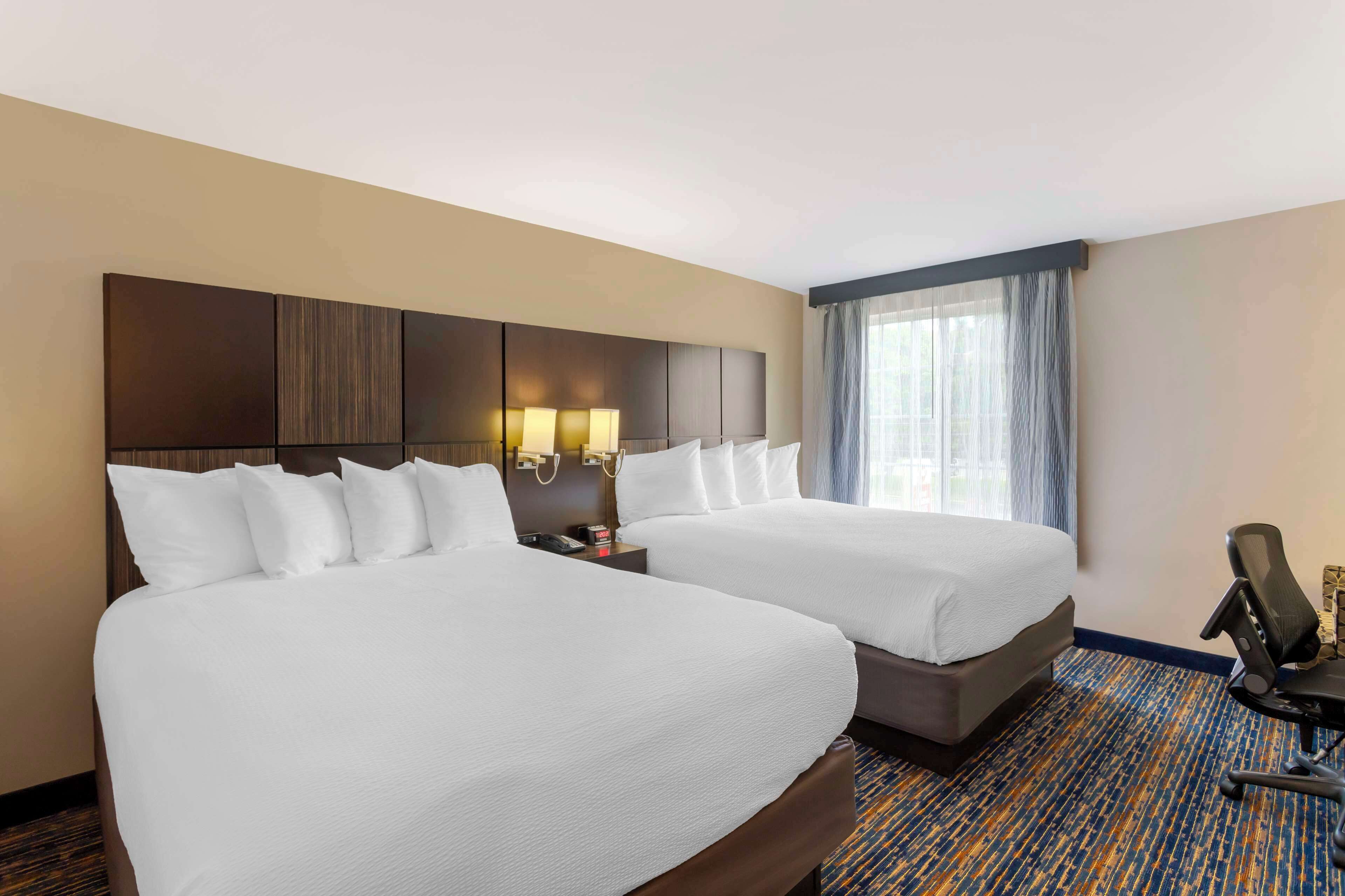 A hotel room with two beds, a desk with a lamp, a chair, and a large window with curtains.