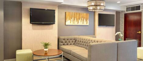 A modern hotel room with a sectional sofa, a flat-screen TV, a chandelier, and a decorative wall piece.