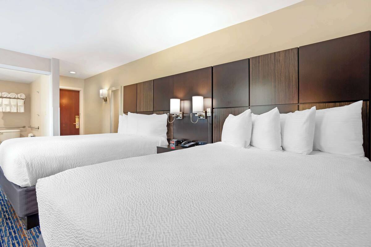 A hotel room with two beds, a wooden headboard, wall-mounted lights, and a bathroom visible through an open door.