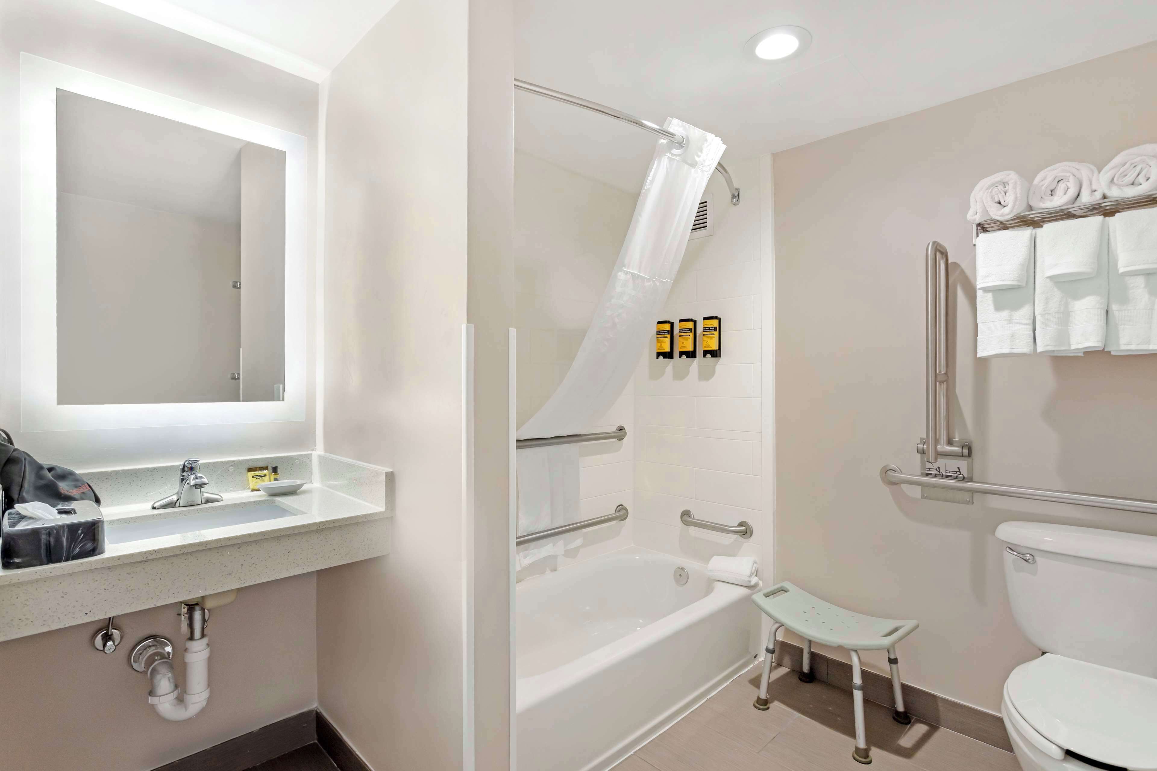 A bathroom with a sink, mirror, shower, toilet, and handrails.