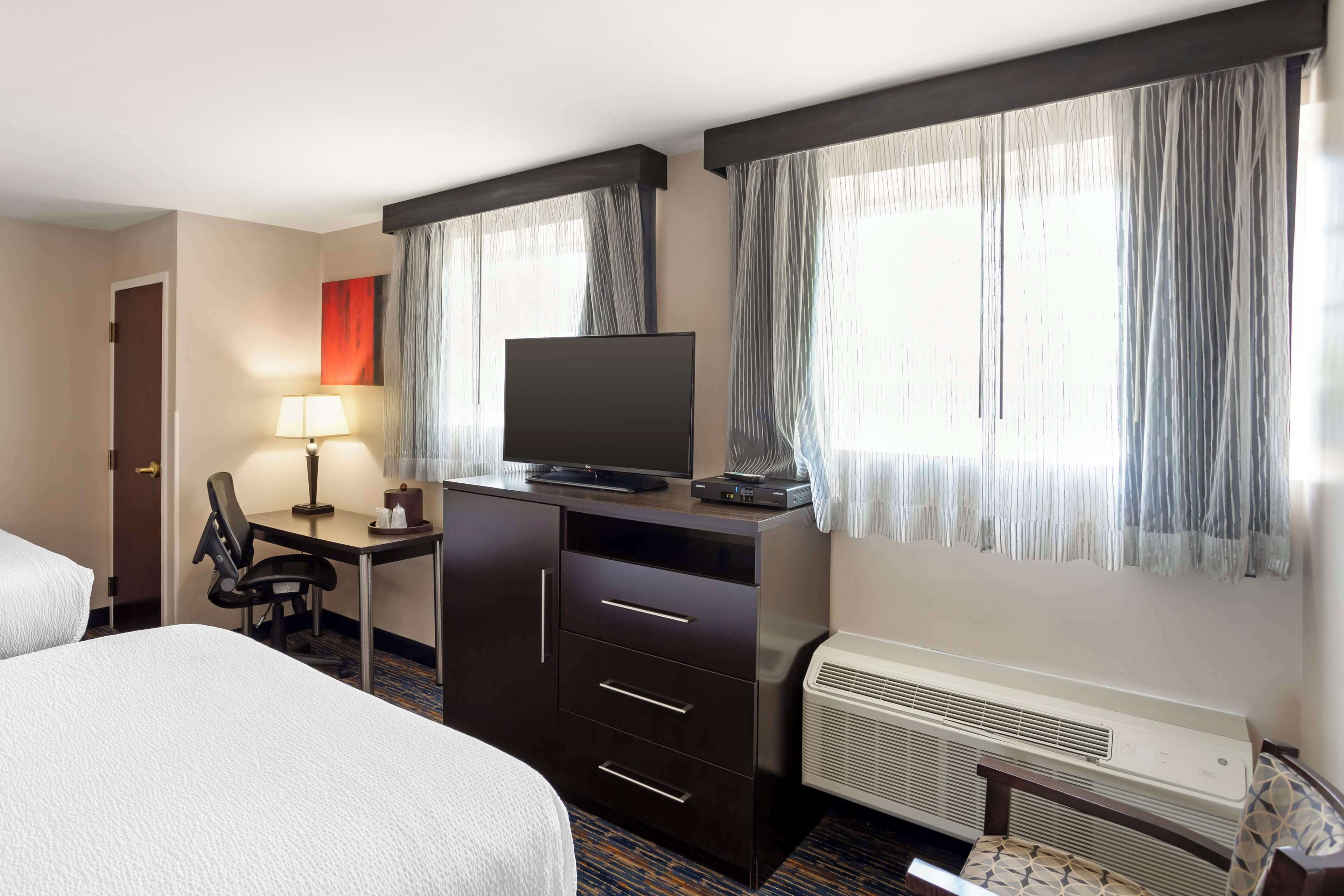 A hotel room with a bed, a desk with a chair, a television, and an air conditioning unit.