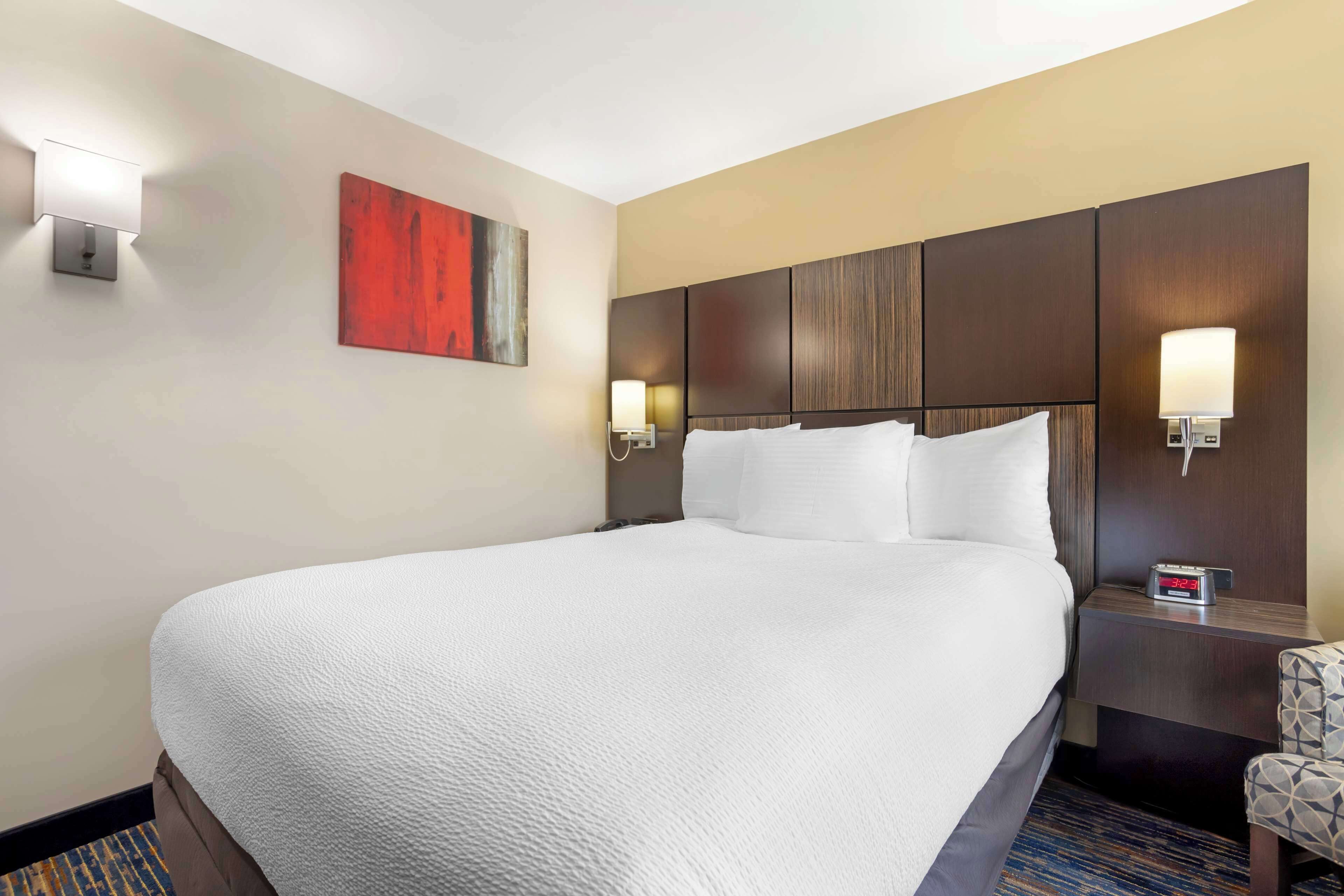 A hotel room with a large bed, a headboard with panels, two wall-mounted lamps, a red abstract painting, and a clock on the nightstand.