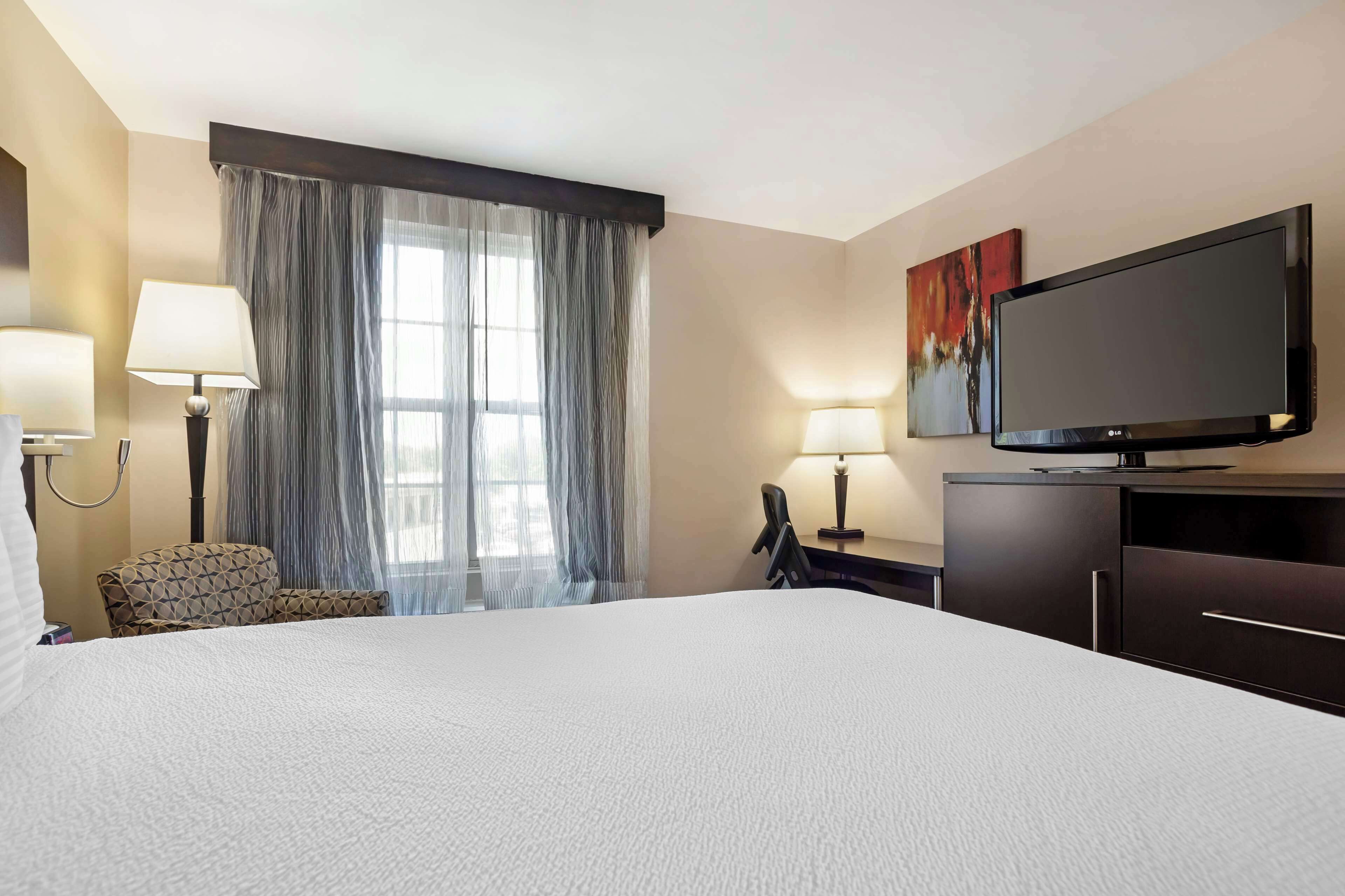 Hotelkamer met een groot bed, een flatscreen televisie, een bureau met stoel, een lamp en een raam met dunne gordijnen.
