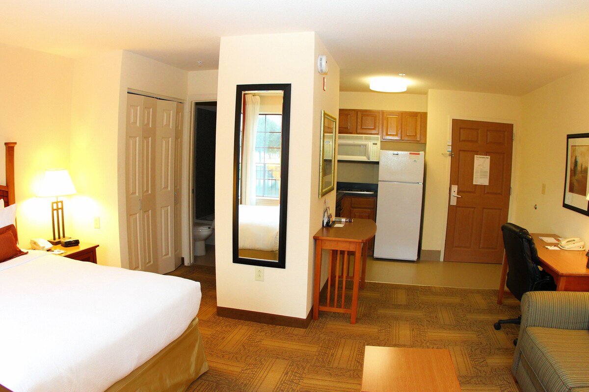 A hotel room with a bed, a desk, a chair, a kitchenette, and a bathroom.
