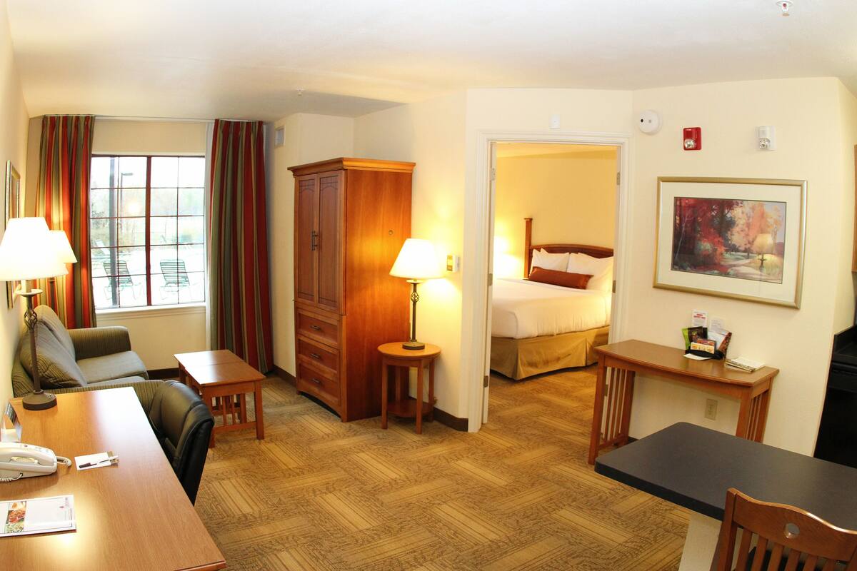 A hotel room with a wooden desk, a chair, a bed, a small table with a lamp, and a painting on the wall.