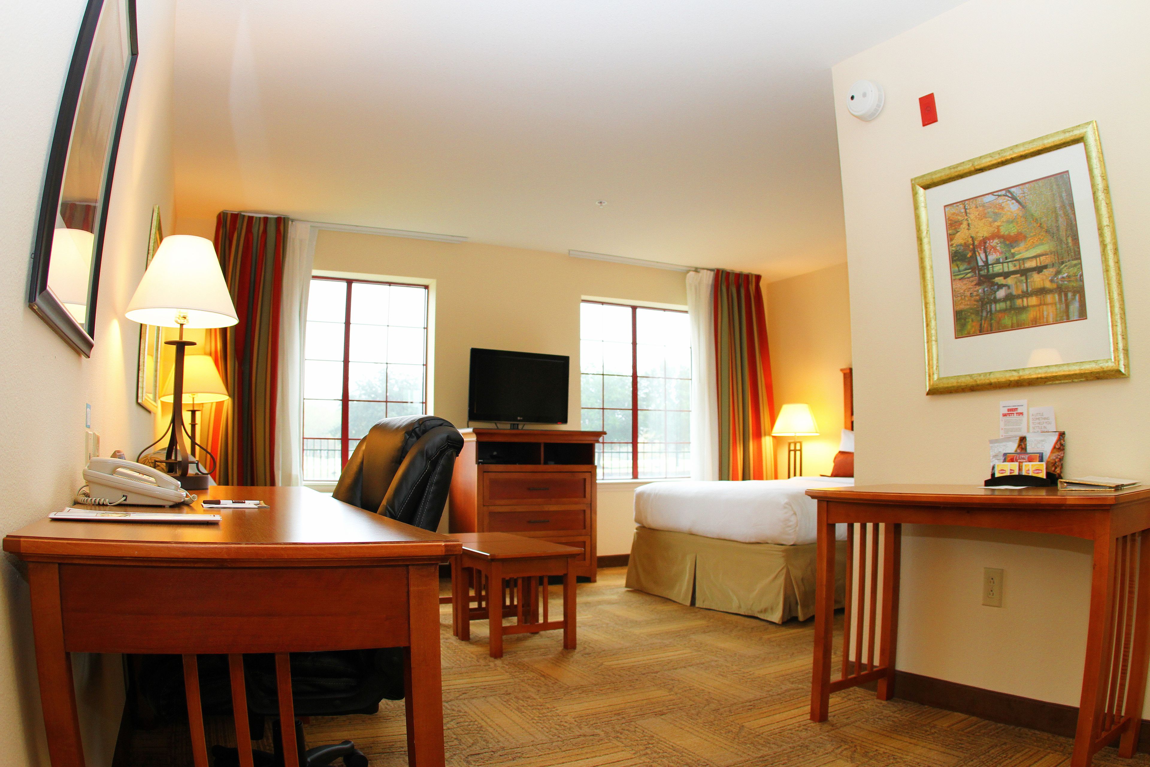 A hotel room with a desk, chair, bed, television, and a painting on the wall.
