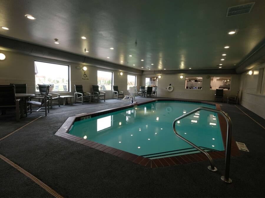 Una piscina interior con suelo de baldosas, rodeada de sillas y mesas, y grandes ventanales que aportan luz natural.