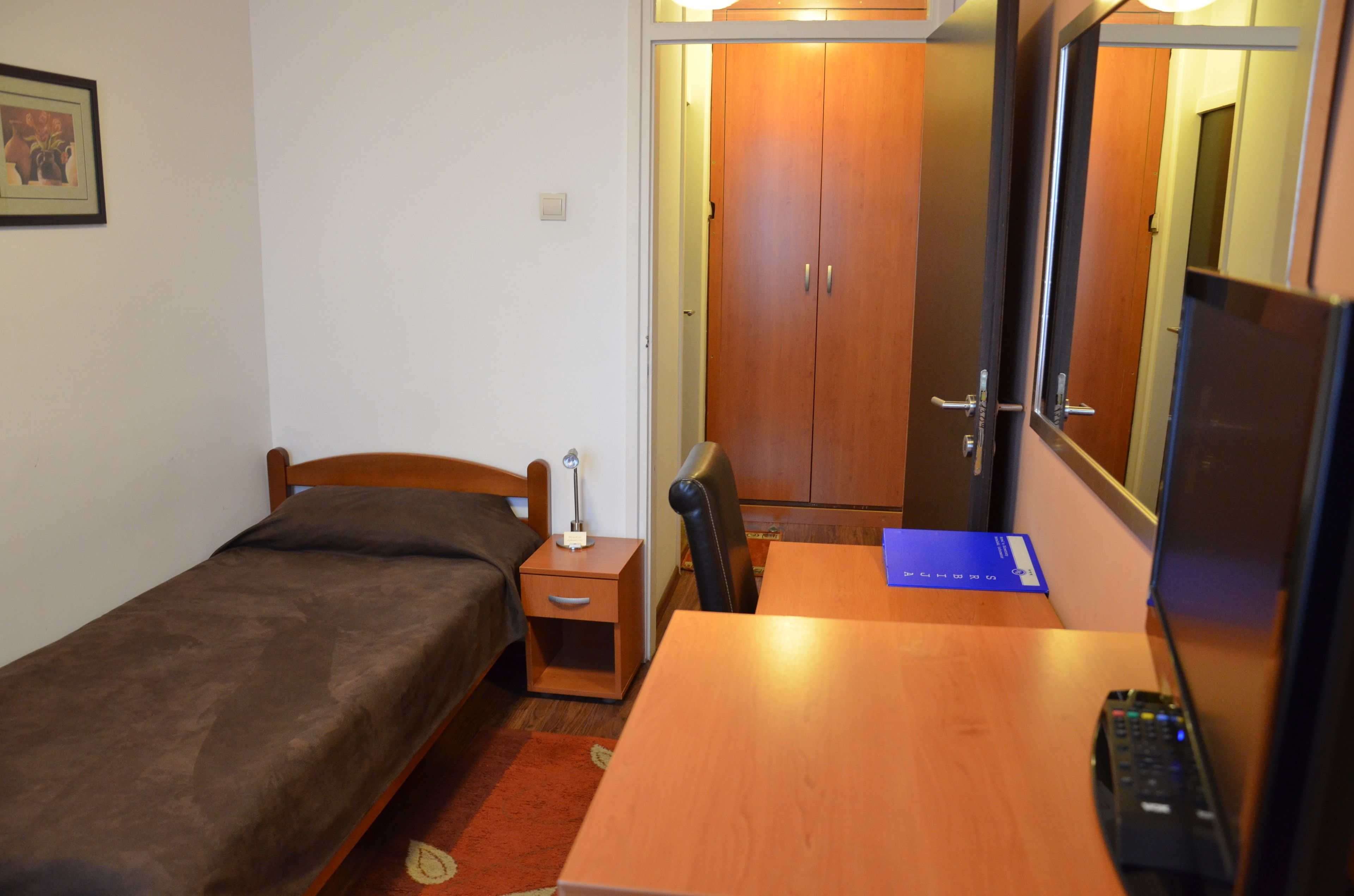Une petite chambre avec un lit simple, un bureau en bois, une chaise, une table de chevet avec une lampe et un téléviseur.