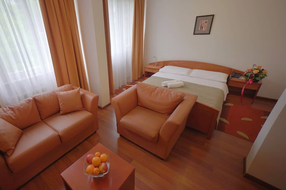 A hotel room with a bed, two sofas, a small table with fruit, and a nightstand with a phone and flowers.