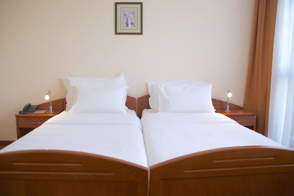A double bed with white linens, two bedside tables with lamps, a framed picture on the wall, and a window with curtains.