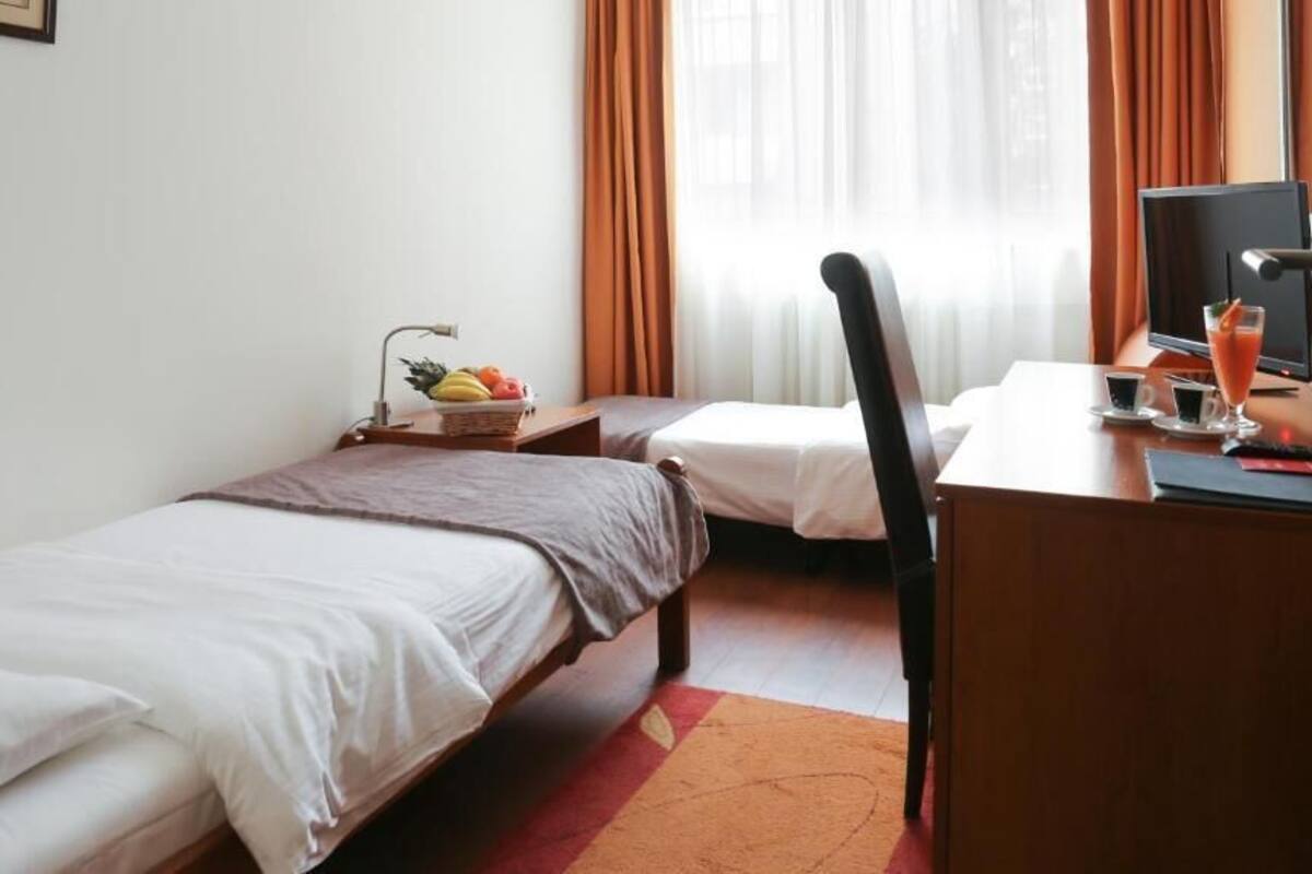 A hotel room with two single beds, a desk with a computer, a small table with a fruit bowl, and a TV.