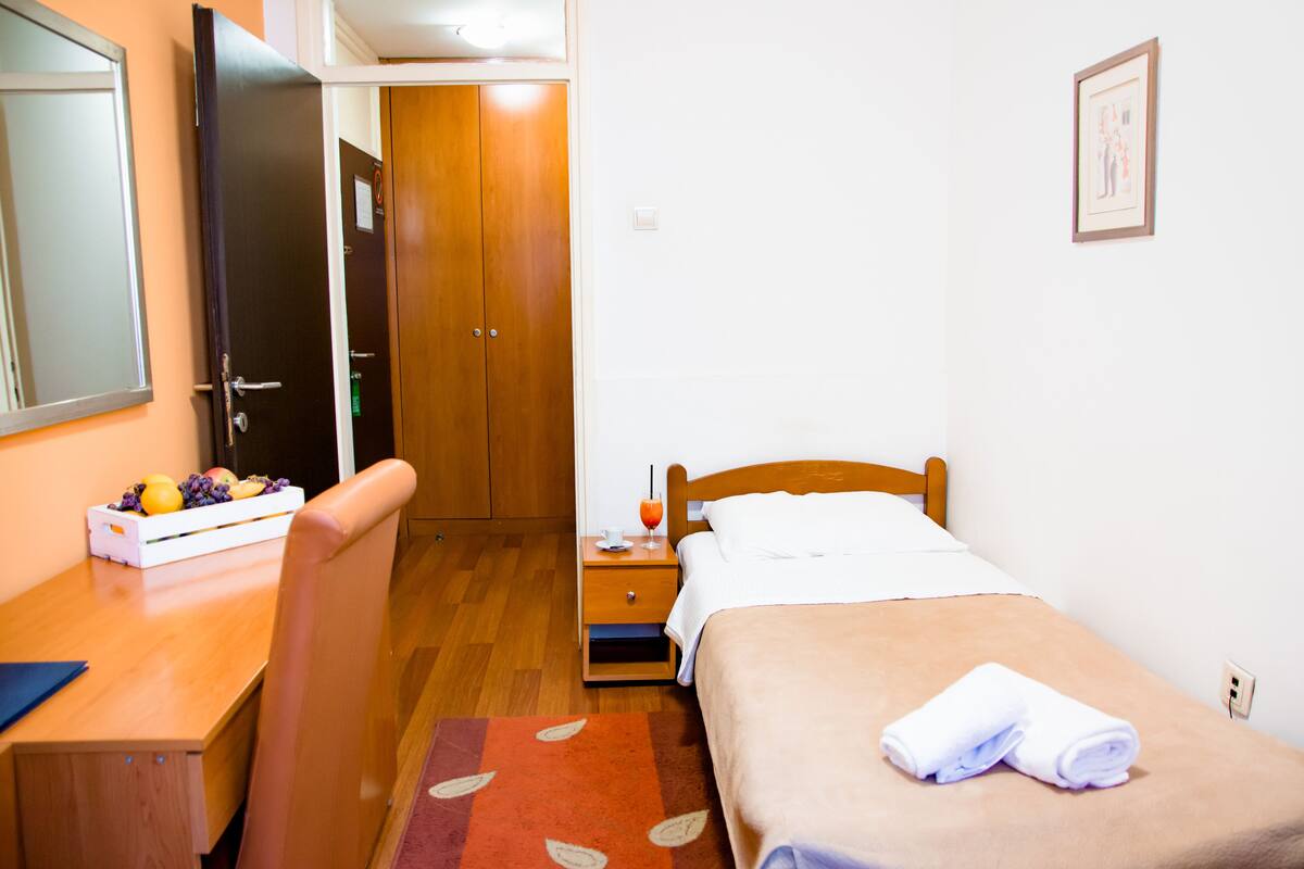 A hotel room with a bed, a wooden nightstand, a desk with a chair, a wardrobe, and a small table with fruit.