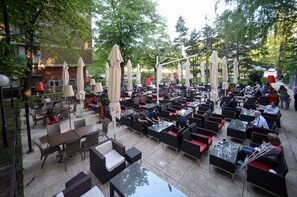 An outdoor cafe with multiple tables and chairs, umbrellas, and a view of the surrounding trees and buildings. - Hotel Srbija (Belgrade)