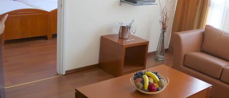 A hotel room with a wooden floor, a bed with white linens, a television on a stand, a small table with a fruit bowl, a glass of orange juice, and a sofa.