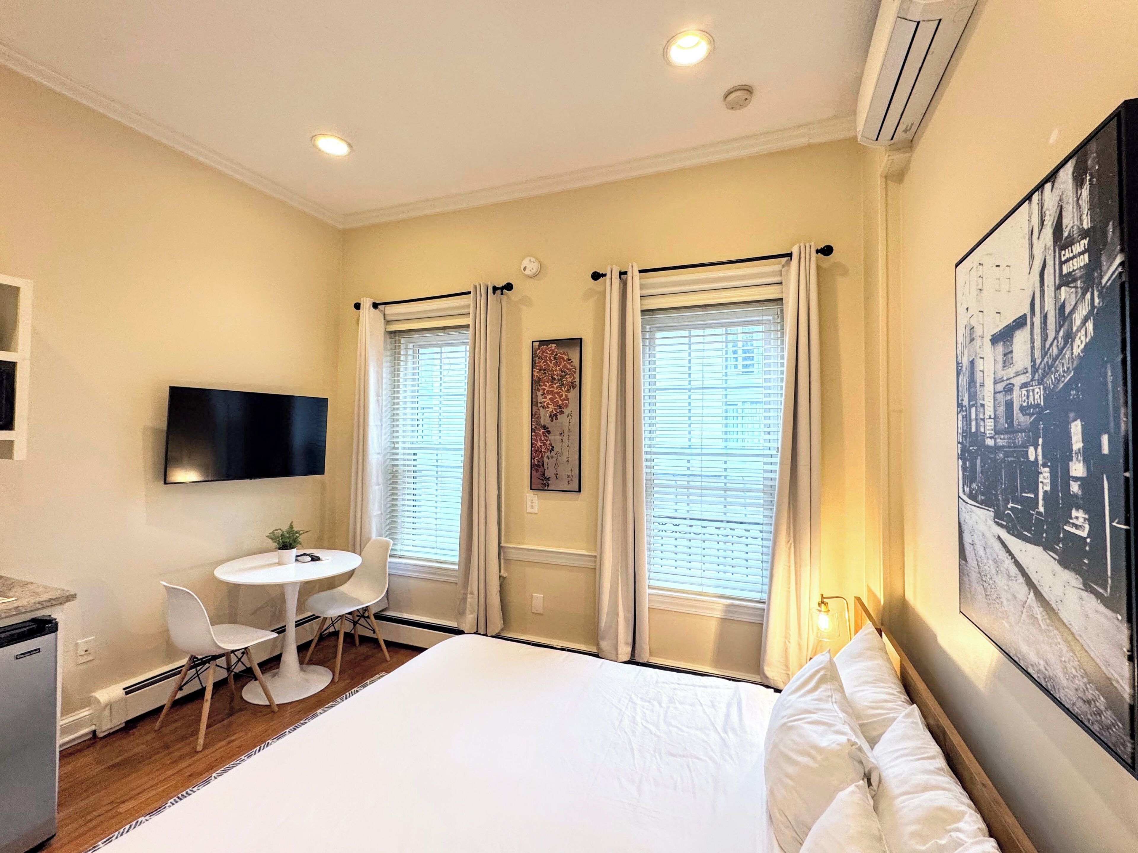 Habitación de hotel con cama, escritorio con silla, televisor en la pared y dos ventanas con cortinas.
