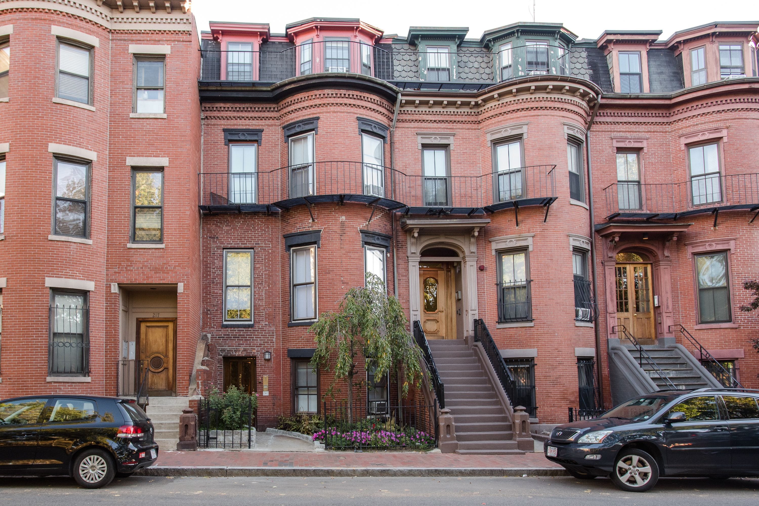 A row of red brick townhouses with multiple stories, each featuring a front door, windows, and a small garden area.