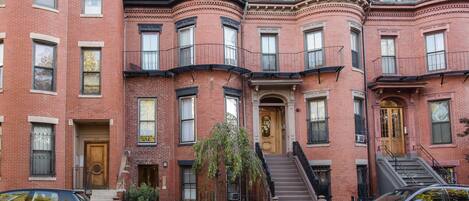 A row of red brick townhouses with multiple stories, each featuring a front door, windows, and a small garden area.