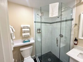 A glass-enclosed shower stall with a white towel hanging on the door, a toilet, and a sink with a soap dispenser.