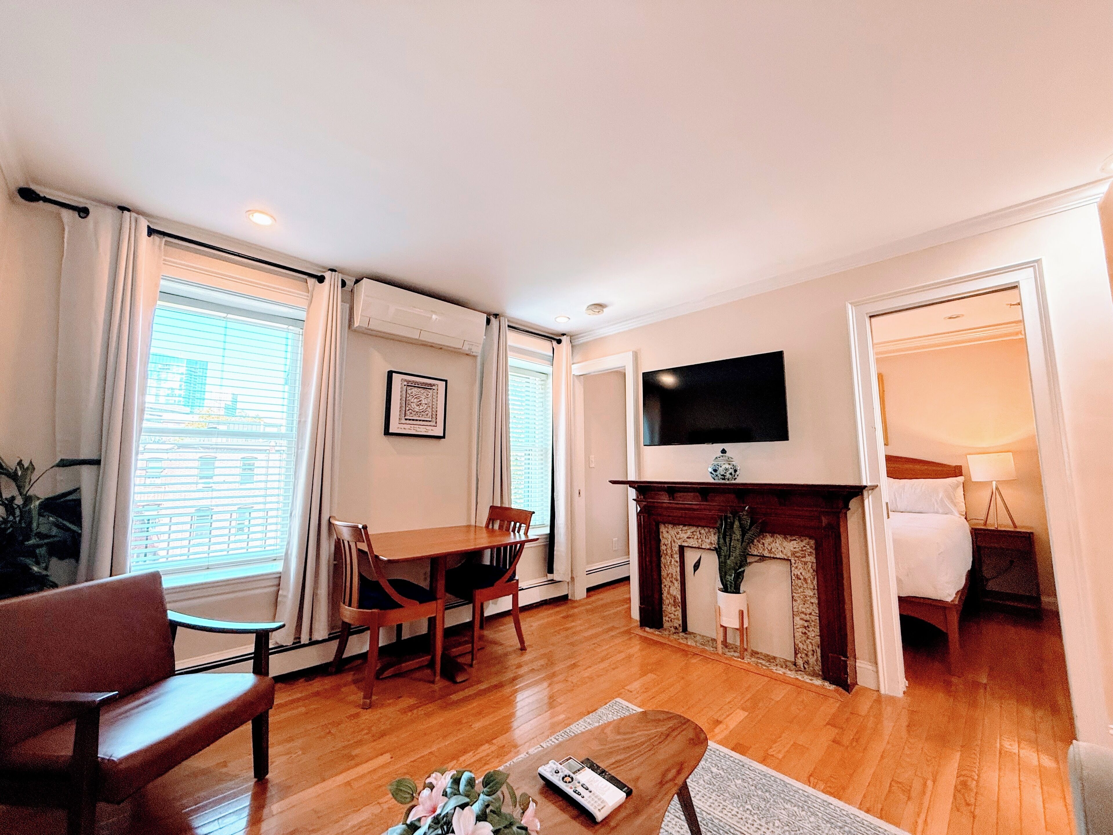 A hotel room with a wooden floor, a dining table with chairs, a flat-screen TV mounted on the wall, a fireplace, and a bed with a nightstand.