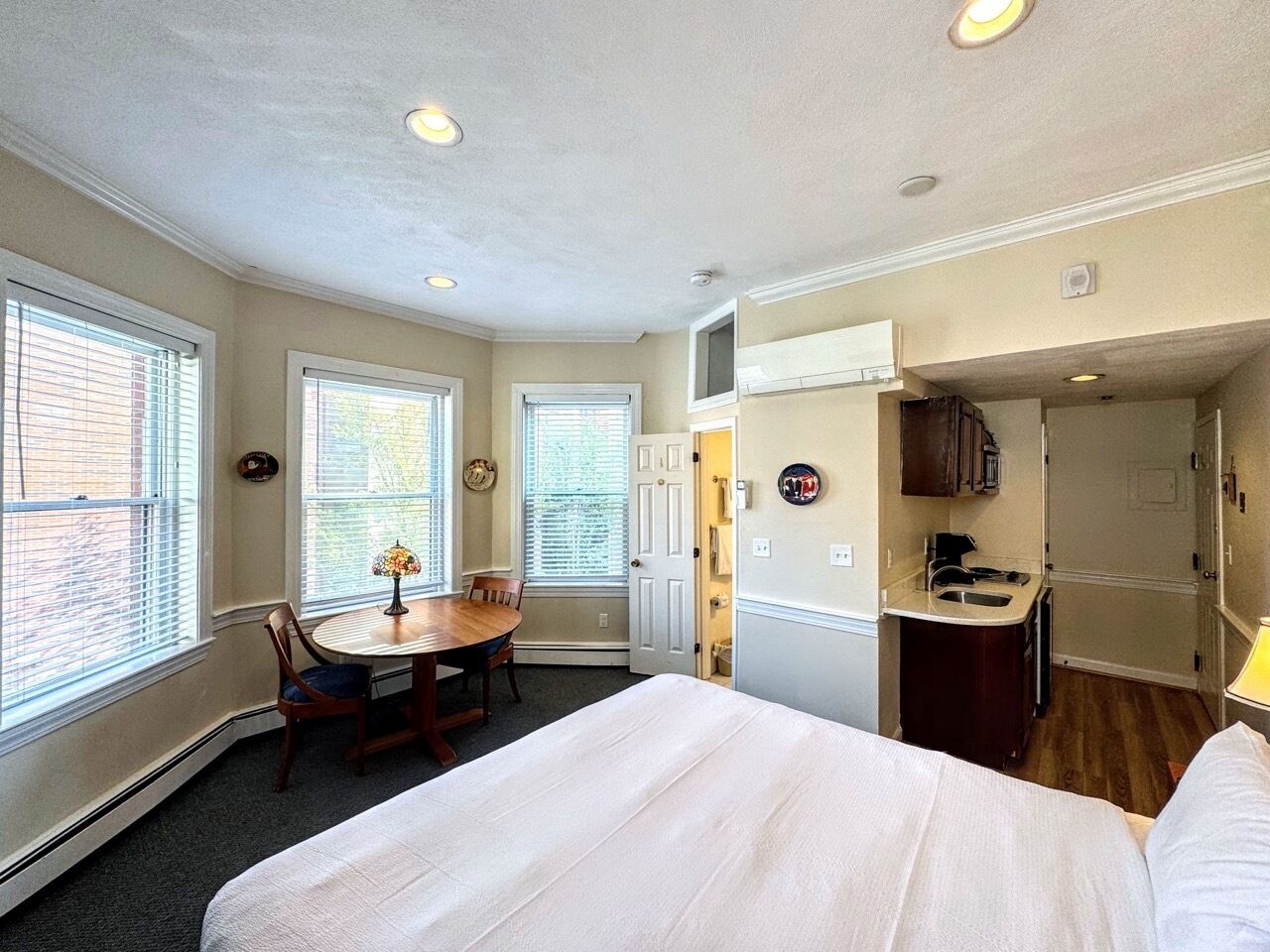 A compact hotel room with a bed, a dining table with chairs, a small kitchen area, and a window with blinds.