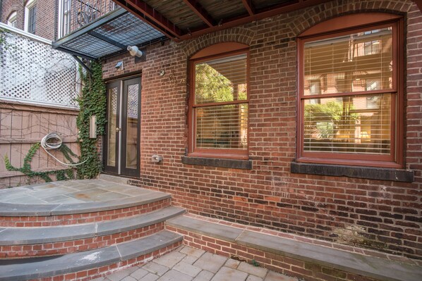 A brick building with a set of steps leading to a door, a window with blinds, and a metal awning above the door.