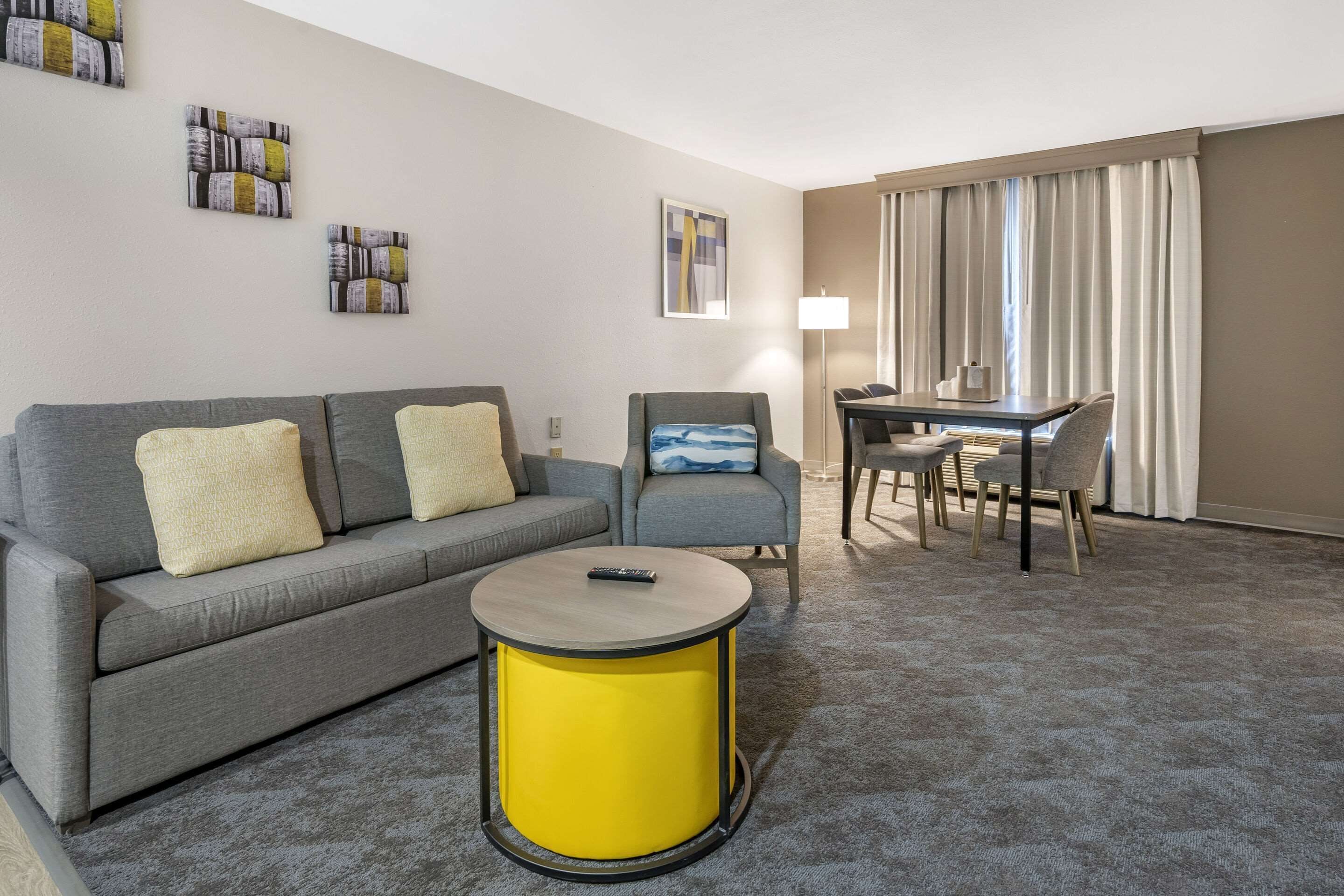 Una sala de estar moderna con un sofá gris, una mesita auxiliar amarilla, un sillón gris y una mesa de comedor con sillas.