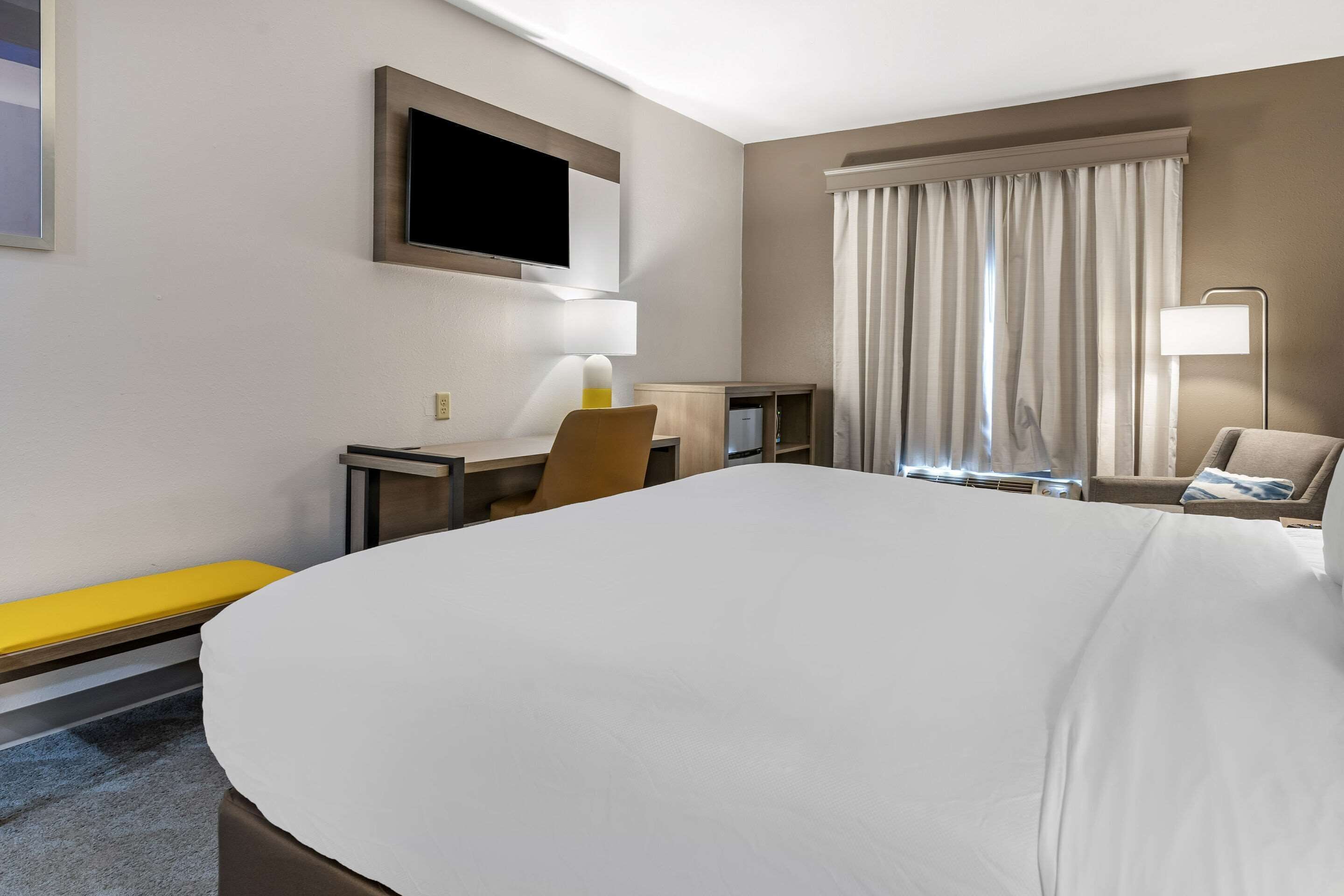 Una habitación de hotel con una cama grande, un escritorio con silla, un televisor montado en la pared y una ventana con cortinas.