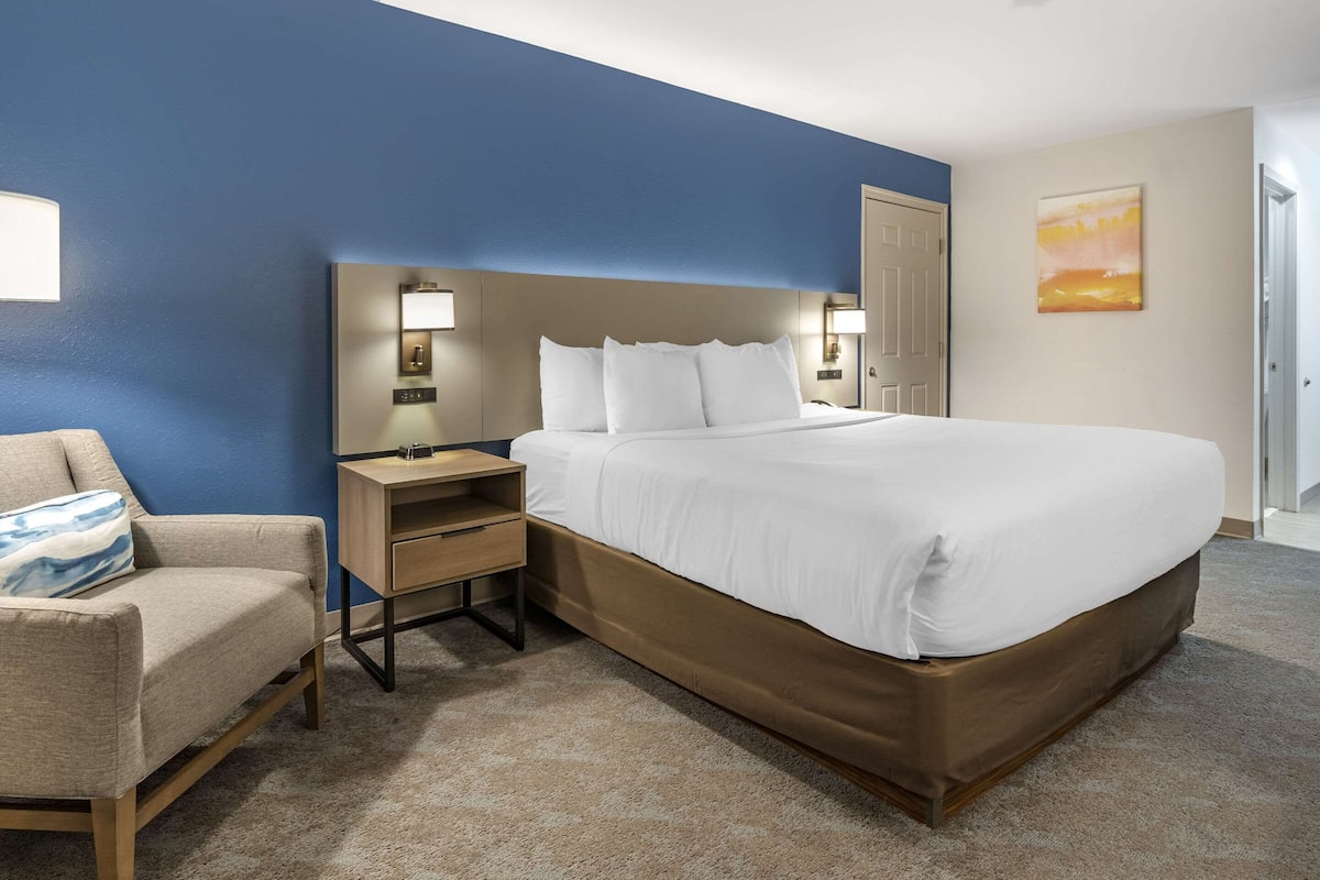A hotel room with a large bed, a bedside table with a lamp, a chair, and a painting on the wall.