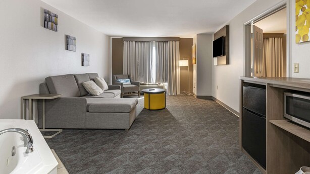 A modern hotel room with a grey sofa, a small round table, a flat-screen TV, and a minibar. - Comfort Inn & Suites Shelbyville (Shelbyville)
