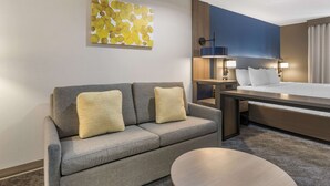 A hotel room with a grey sofa, a round table, a bed with white linens, and a wall-mounted abstract art piece. - Comfort Inn & Suites Shelbyville (Shelbyville)