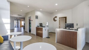A modern reception area with a wooden reception desk, a computer, and a phone. There are three white round tables and a grey sofa with a yellow pillow. - Comfort Inn & Suites Shelbyville (Shelbyville)