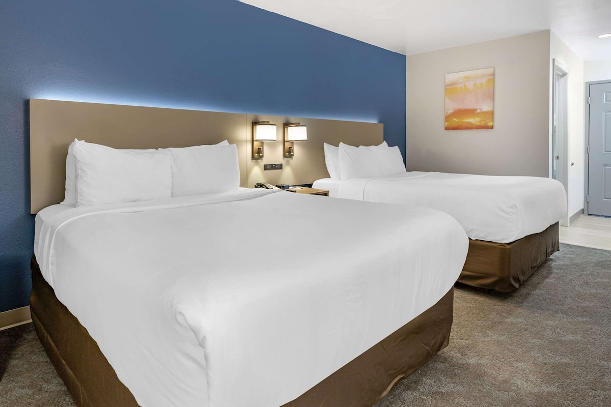 A hotel room with two beds, a headboard with integrated lighting, a nightstand with a phone, and a painting on the wall.