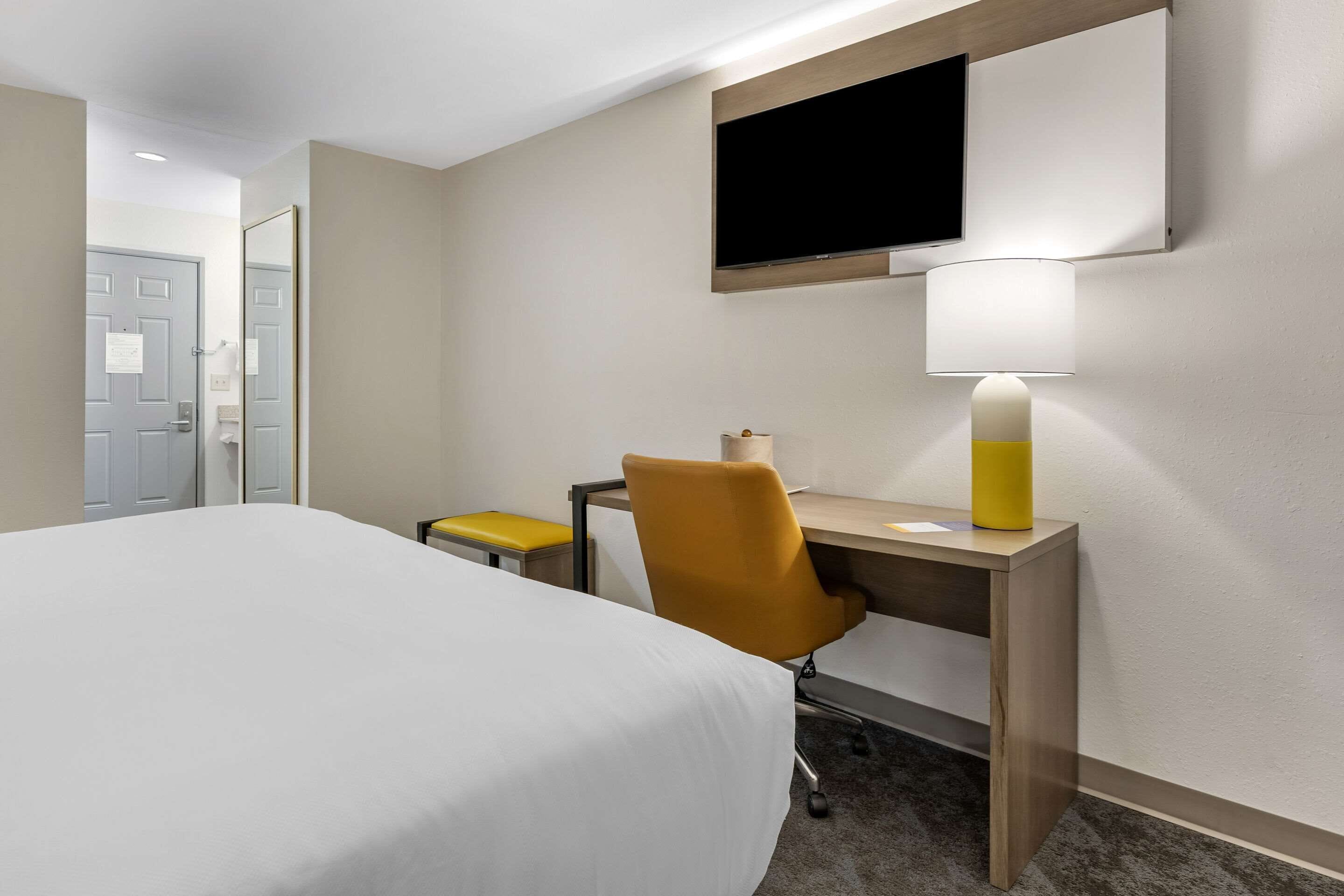 A hotel room with a bed, a desk with a chair, a television mounted on the wall, and a lamp on the desk.