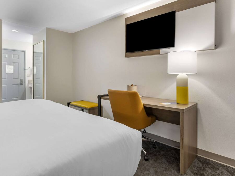 Quarto de hotel com uma cama, uma escrivaninha com cadeira, uma televisão na parede e um abajur na escrivaninha.
