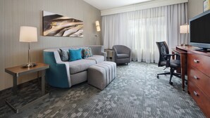 A hotel room with a grey sectional sofa, a small side table, a desk with a chair, a television, and a large window with curtains. - Courtyard by Marriott Grand Junction (Grand Junction)