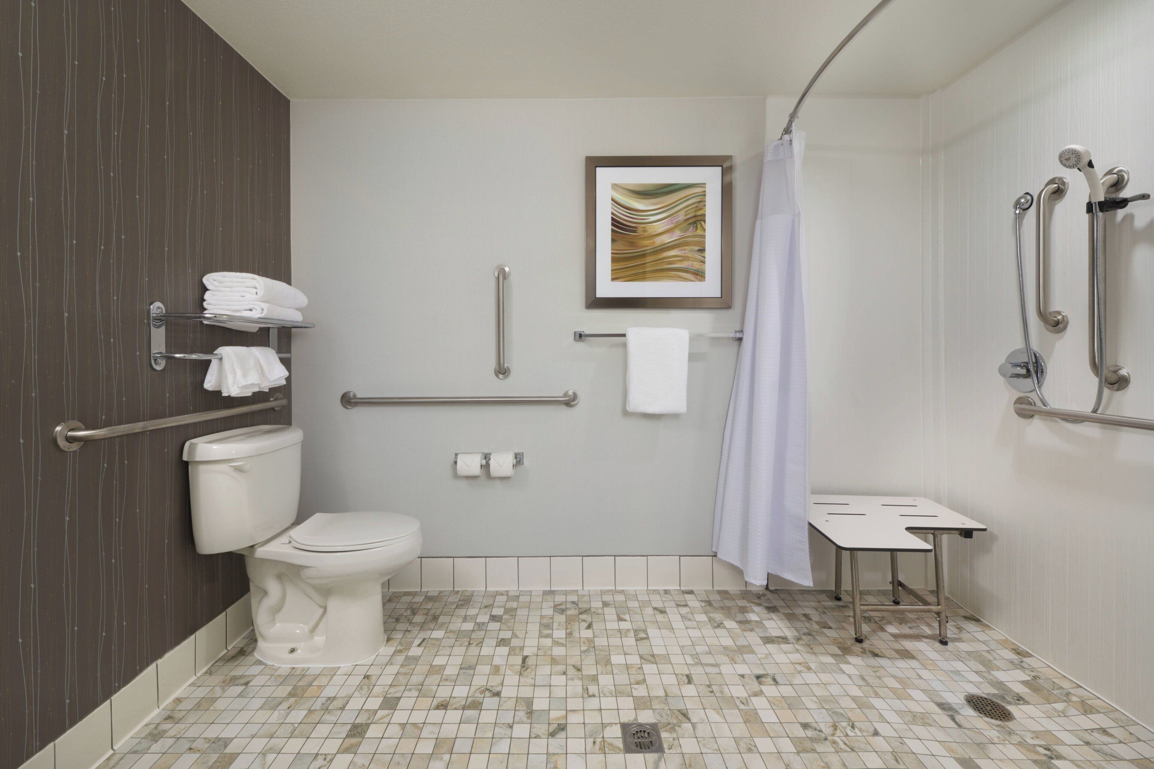 A bathroom with a toilet, grab bars, a shower with a handheld showerhead, a towel rack, and a small table.