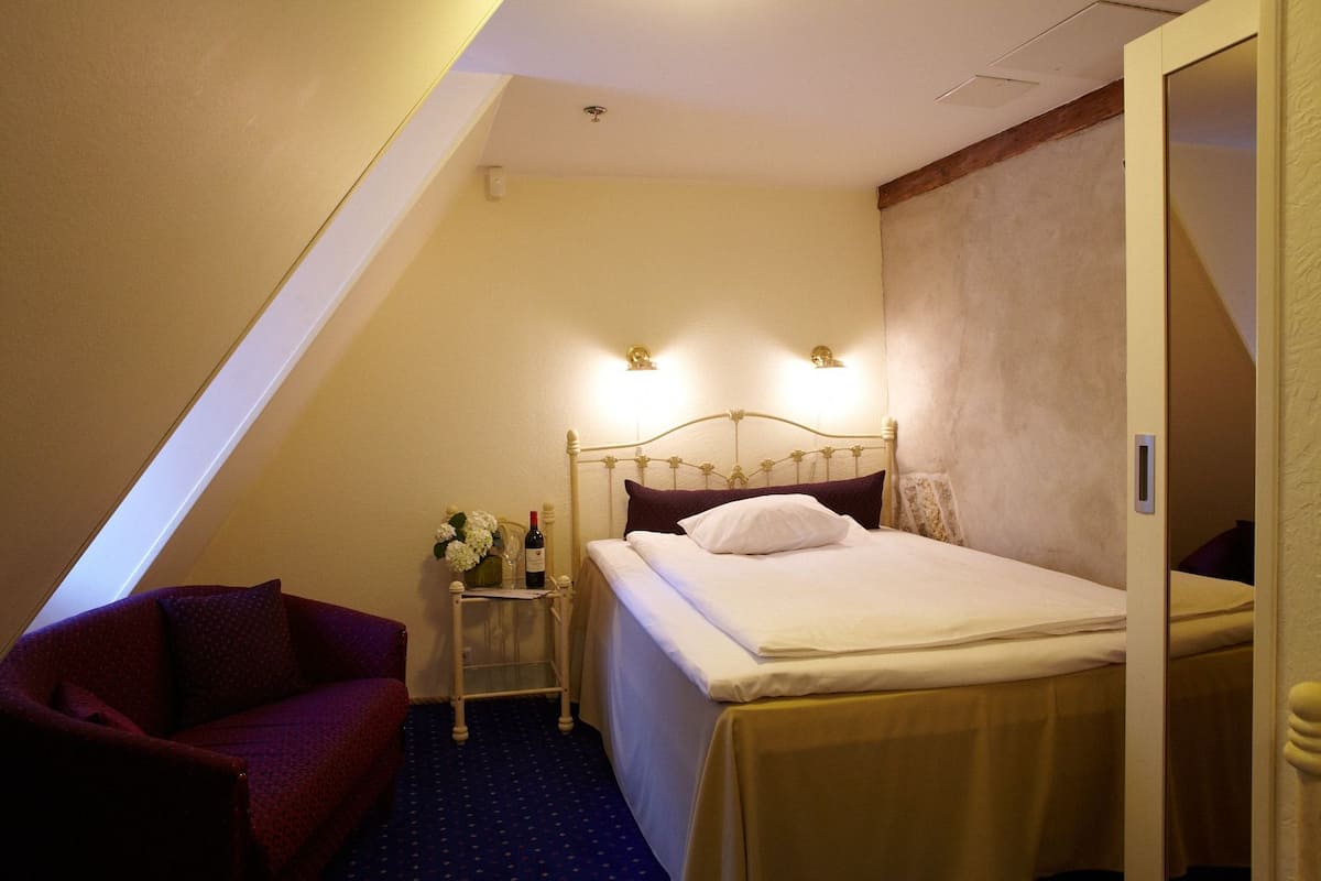 A small, cozy hotel room with a single bed, a nightstand, a chair, and a small table with a vase and wine bottle.