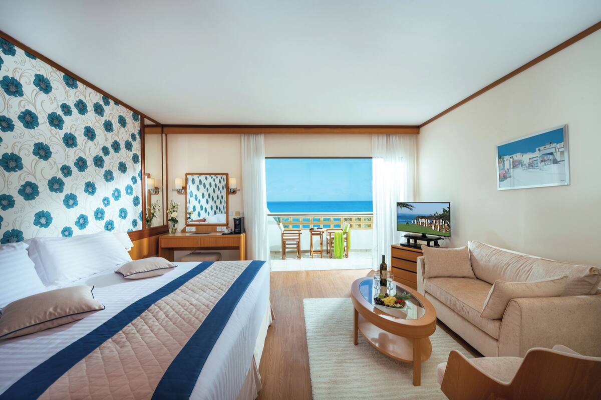 Une chambre d’hôtel avec un lit, un canapé, une télévision, un bureau et un balcon donnant sur la mer.