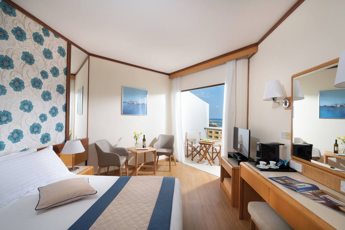 Une chambre d’hôtel avec un lit, un bureau avec un ordinateur, une chaise, une petite table avec un vase et une fenêtre donnant sur la mer.
