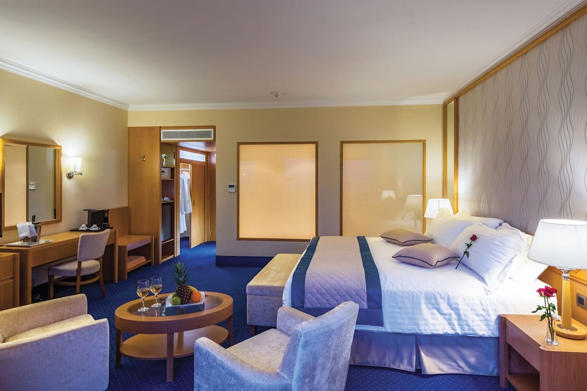 Une chambre d’hôtel comprenant un lit, un bureau, une chaise, une petite table avec des fruits et des boissons, ainsi qu’une salle de bain avec douche et toilettes.