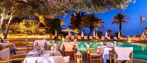 Un área de comedor al aire libre con mesas dispuestas para una comida, con vista a una piscina y palmeras.
