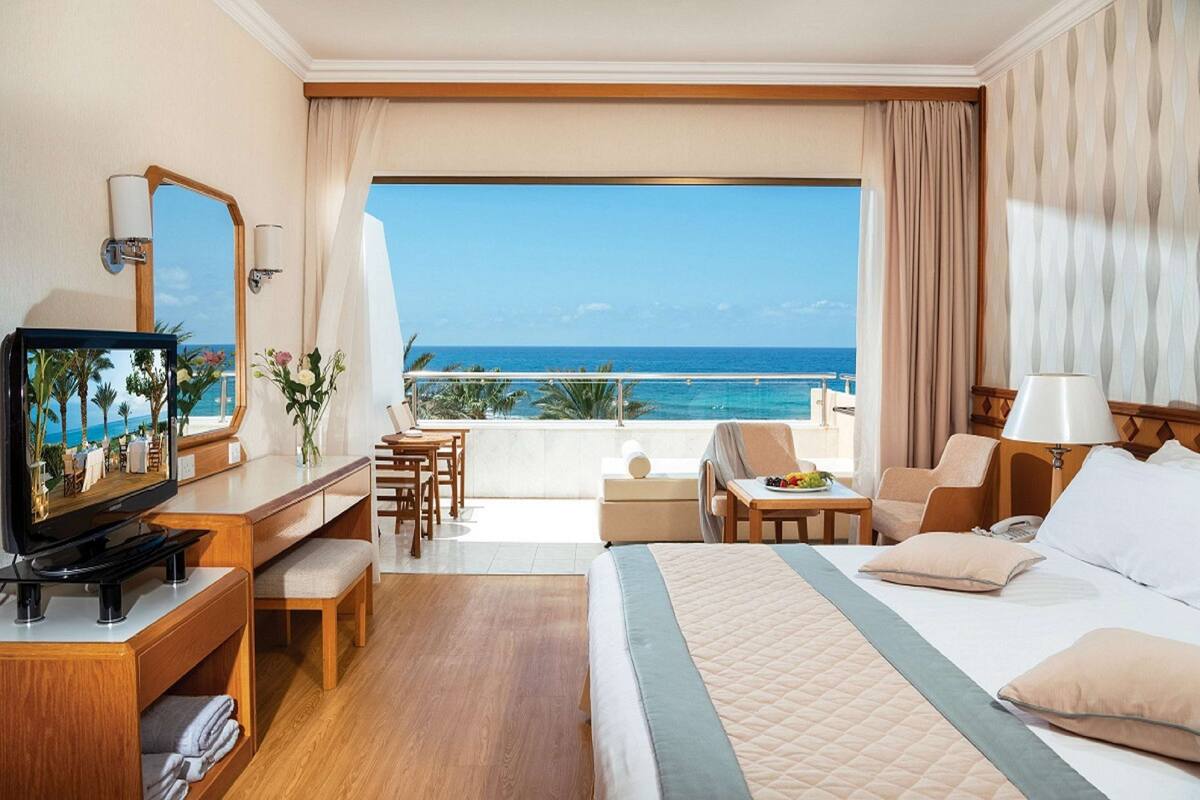 Ein Hotelzimmer mit einem großen Bett, einem Schreibtisch mit Fernseher, einem Balkon mit Meerblick und einem Essbereich.