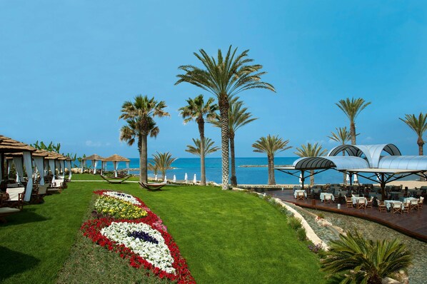 Un resort frente a la playa con mesas al aire libre, palmeras y un jardín de flores.