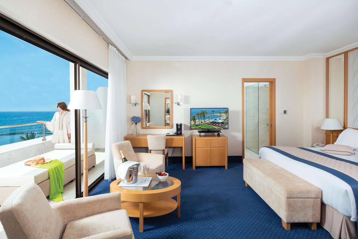 Ein Hotelzimmer mit großem Fenster, einem Bett, einem Schreibtisch, einem Sessel, einem kleinen Tisch und einem Fernseher.