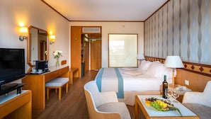 Ein Hotelzimmer mit einem Bett, einem Schreibtisch mit Fernseher, einem Sessel und einem Nachttisch mit einer Obstplatte, sowie einem großen Fenster.