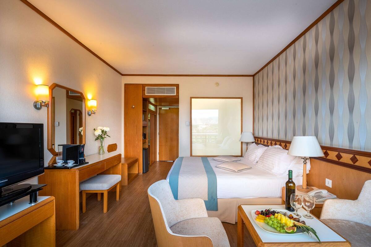 Ein Hotelzimmer mit einem Bett, einem Schreibtisch mit Fernseher, einem Sessel und einem Nachttisch mit einer Obstplatte, sowie einem großen Fenster.