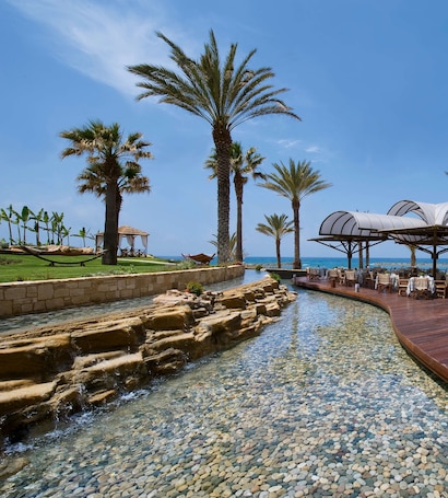 A serene resort setting with a clear pool, palm trees, and a dining area overlooking the sea.