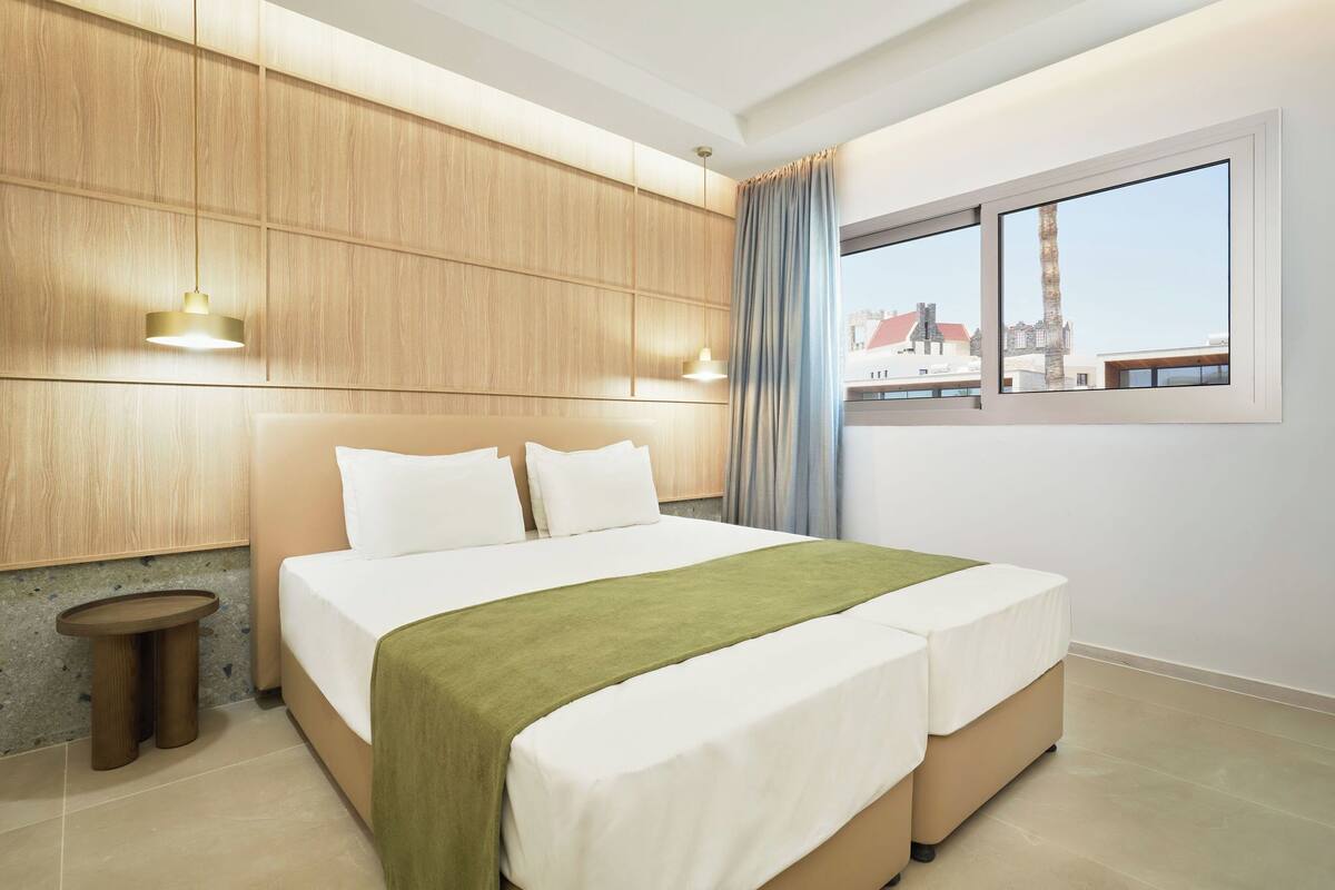 A modern hotel room with a large bed, wooden panel walls, a small side table, and a window with a city view.