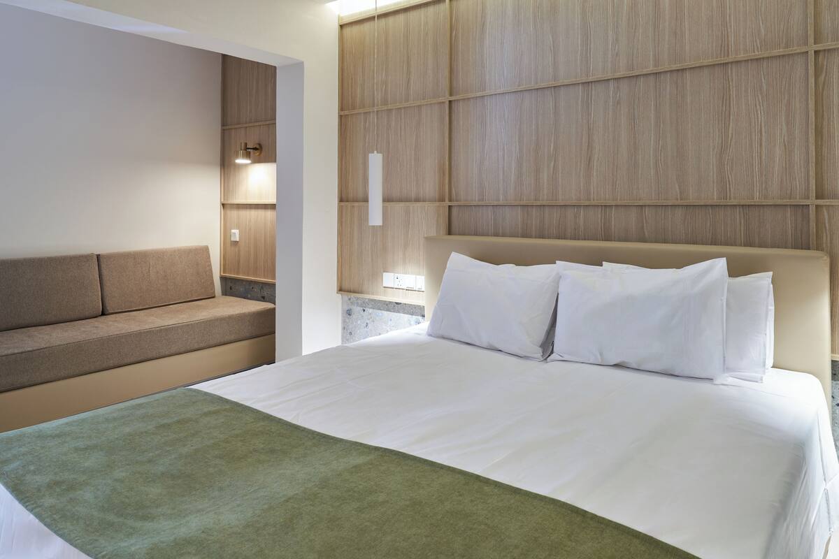A modern hotel room with a large bed, a sofa, and a wall-mounted light fixture.